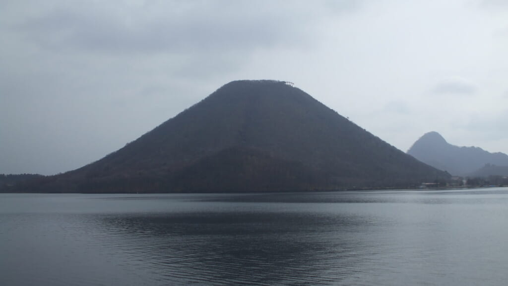 Conical peak of Haruna Fuji rising behind the still water of Lake Haruna under grey cloud, Gunma