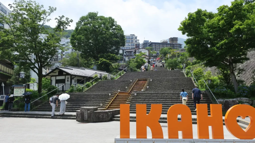 Wide stone stairway climbing between trees and buildings at Ikaho Onsen, with an orange IKAHO sign in the foreground