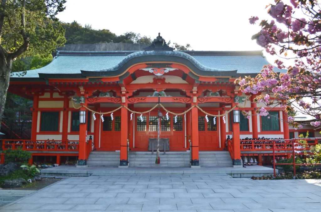 The vermilion hall of Awashima Shrine in Kada, with rows of dolls along its railings and pink cherry blossom at one side