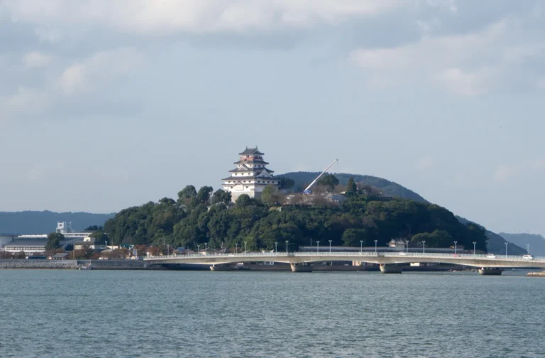 The white keep of Karatsu Castle on a wooded hill above calm water, with a long low bridge in front and hills behind