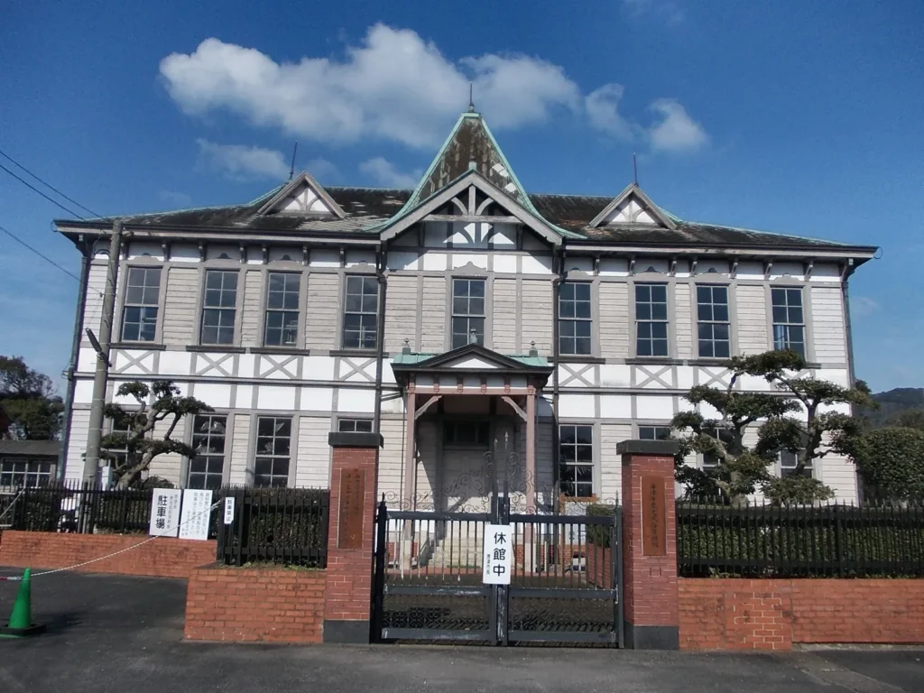 The two-storey white wooden front of the former Mitsubishi Goshi Kaisha Karatsu Branch, with a pointed central gable, behind a brick and iron gate