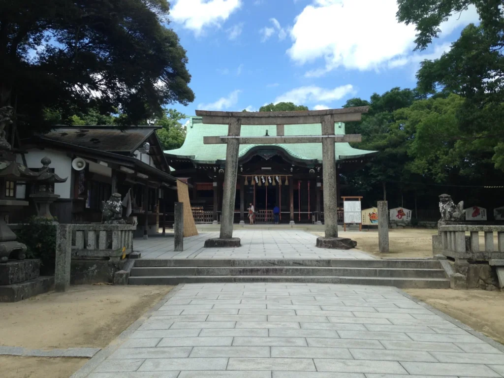 A stone torii on a paved approach leading to the green-roofed worship hall of Karatsu Shrine, with trees around