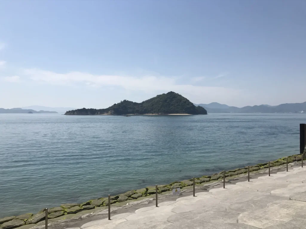 A small wooded island across calm blue water, seen from the stone shore of Okunoshima