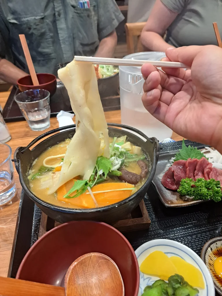 A wide flat hoto noodle lifted from an iron pot of miso broth with pumpkin, carrot and greens