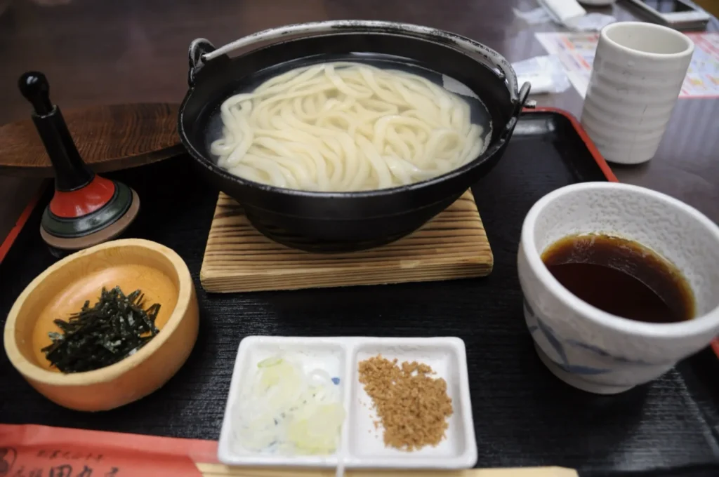 Mizusawa udon served in an iron pot of hot cooking water, with a cup of dipping sauce and side condiments