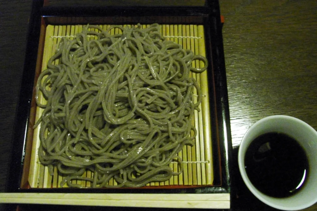 A basket of cold soba noodles on a bamboo mat with a cup of dipping sauce