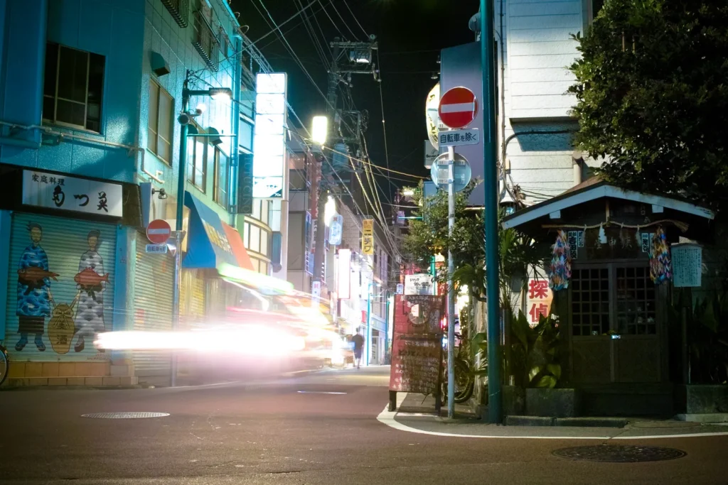 A back street corner in Shimokitazawa at night with shop signs and a small roadside shrine