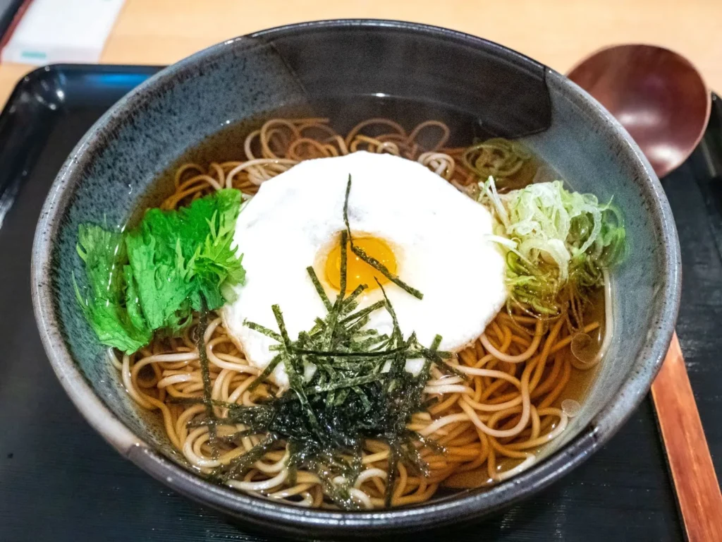 A bowl of hot soba in broth topped with grated yam, an egg yolk, shredded nori and spring onion