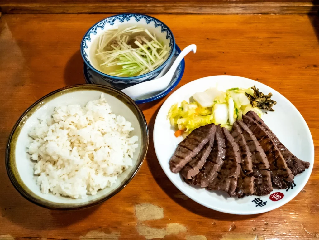 A plate of grilled beef tongue slices with pickles, a bowl of barley rice and a bowl of clear soup