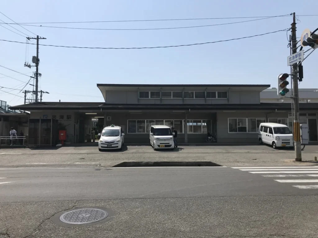 The low building of JR Tadanoumi Station seen across the road, with cars parked in front