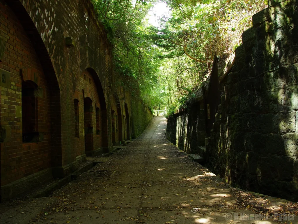 A paved path running between moss-covered brick arches and a stone wall, under a canopy of trees on Tomogashima