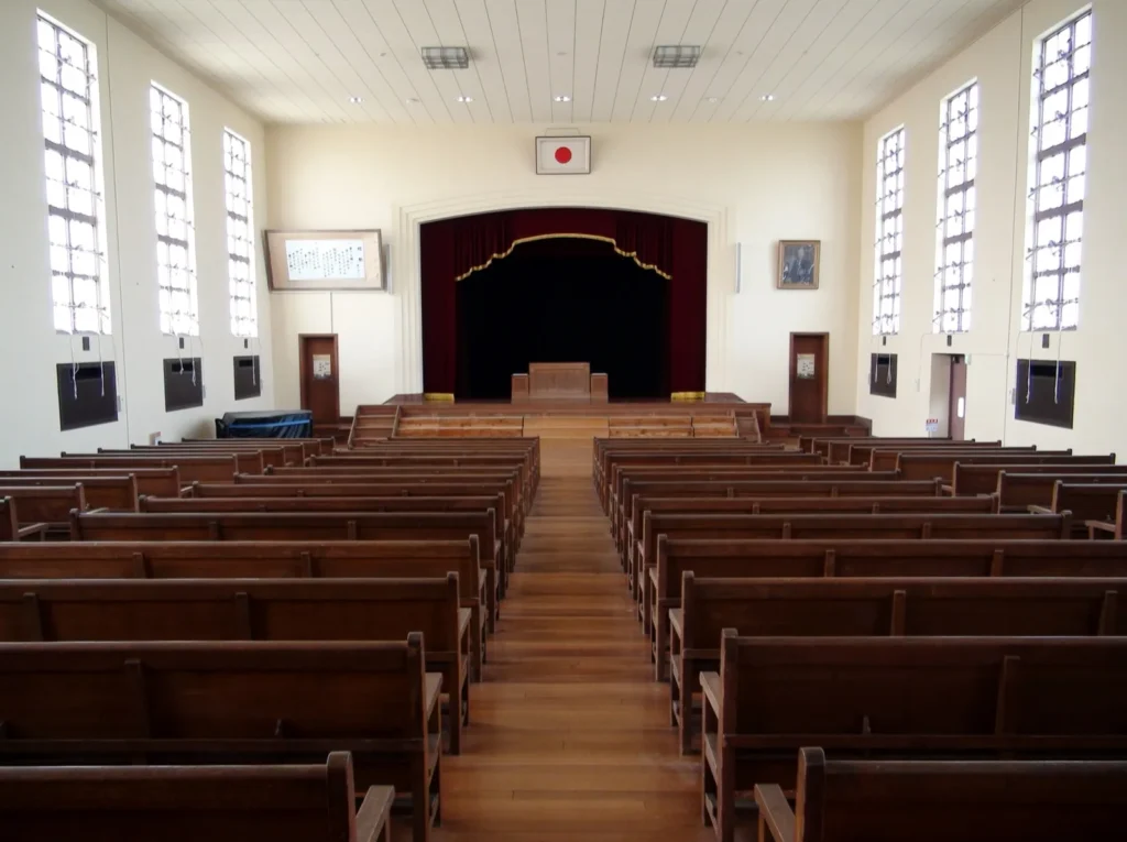 Rows of wooden benches facing the curtained stage of the old school auditorium, with tall windows on both sides