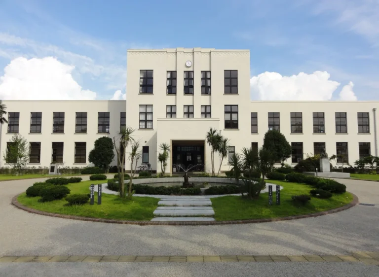 The white three-storey central tower and two-storey wings of the old Toyosato Elementary School behind a round lawn and fountain, under a blue sky